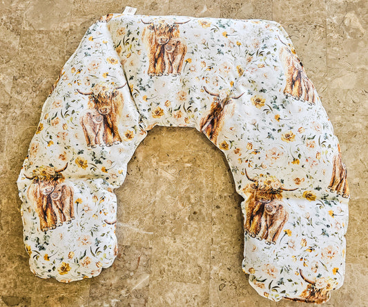 Hot and Cold Neck Pack with a white background, featuring a print of Highland cows and yellow flowers, laid flat on a textured beige stone surface.