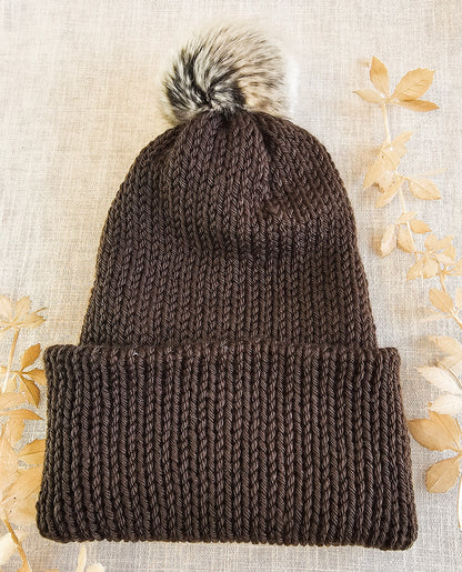Brown knitted beanie with a fur pom-pom on a textured beige background