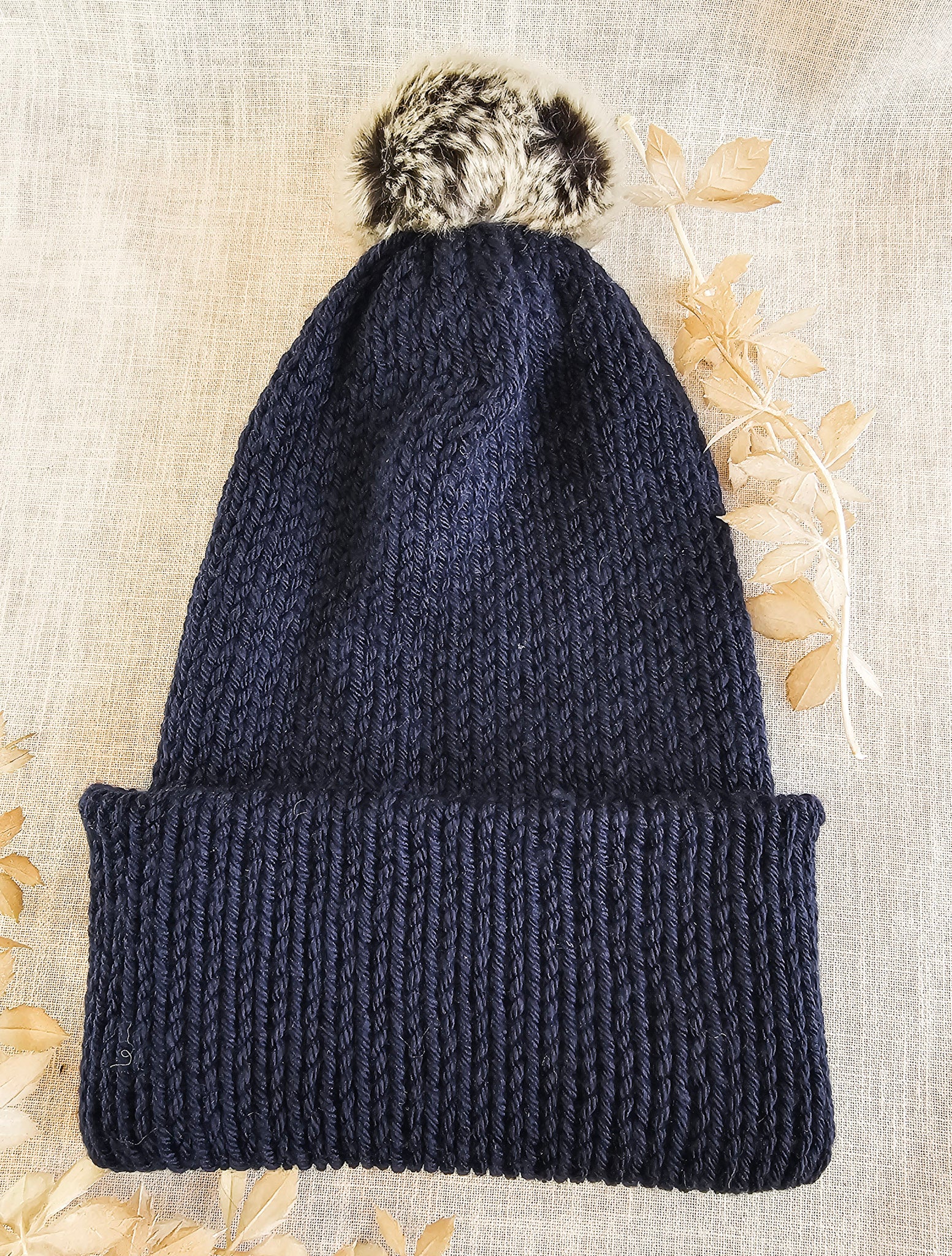 Navy blue knit beanie with a fluffy pom-pom on a textured beige background