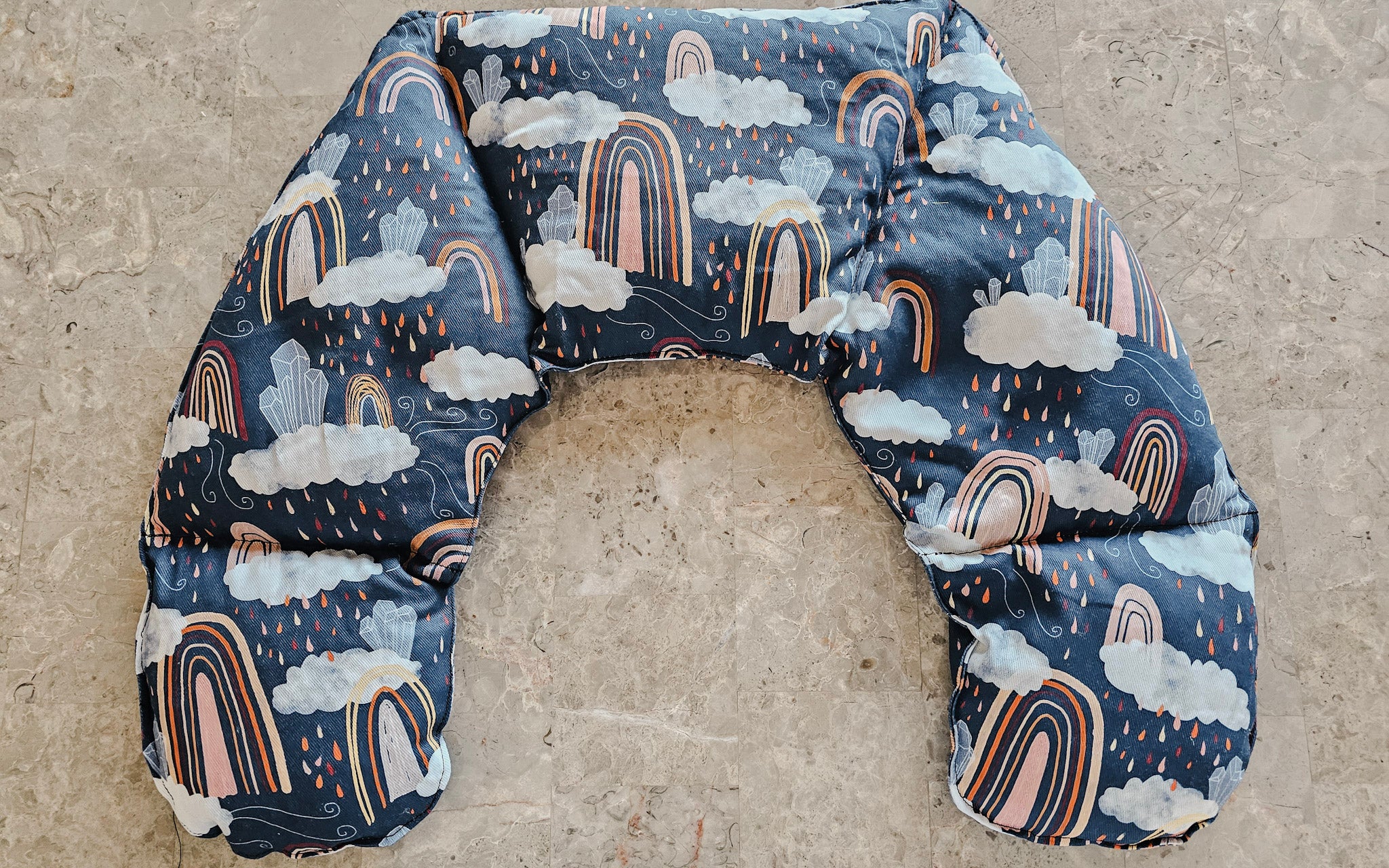 Hot and Cold Neck Pack with a navy blue background, featuring a whimsical print of white clouds, raindrops, and pastel rainbow arches, laid flat on a textured concrete surface.