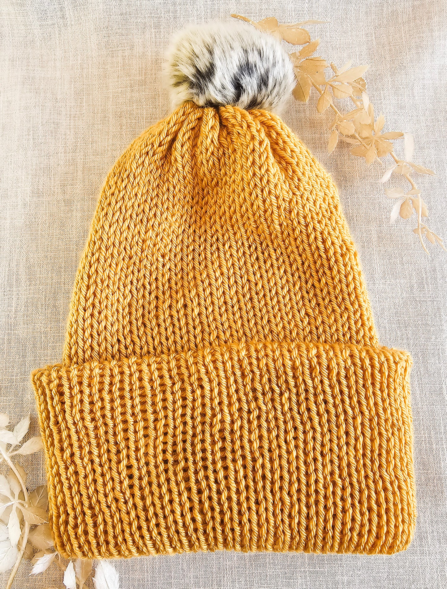 Knitted mustard yellow beanie with a fur pom-pom on a textured beige background