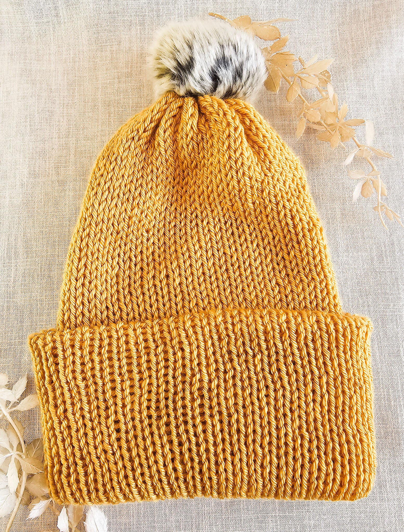 Knitted mustard yellow beanie with a fur pom-pom on a textured beige background