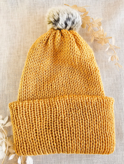 Knitted mustard yellow beanie with a fur pom-pom on a textured beige background