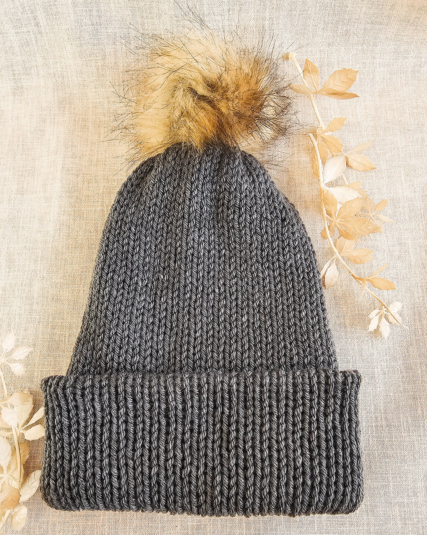 Gray knit beanie with a fur pom-pom on a textured beige background