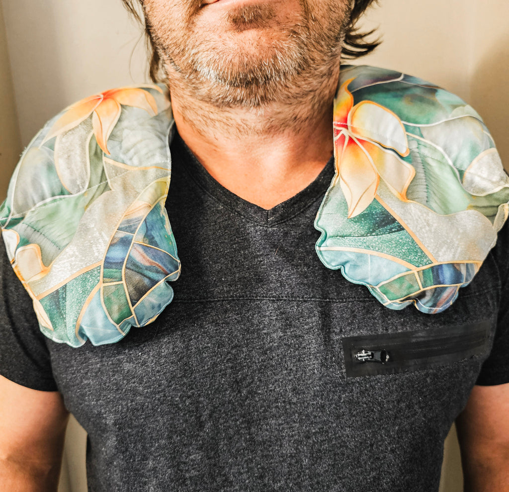 A man wearing a hot and cold neck pack with a tropical floral print in shades of teal, green, yellow, and orange, resting against his shoulders over a dark gray v-neck t-shirt.