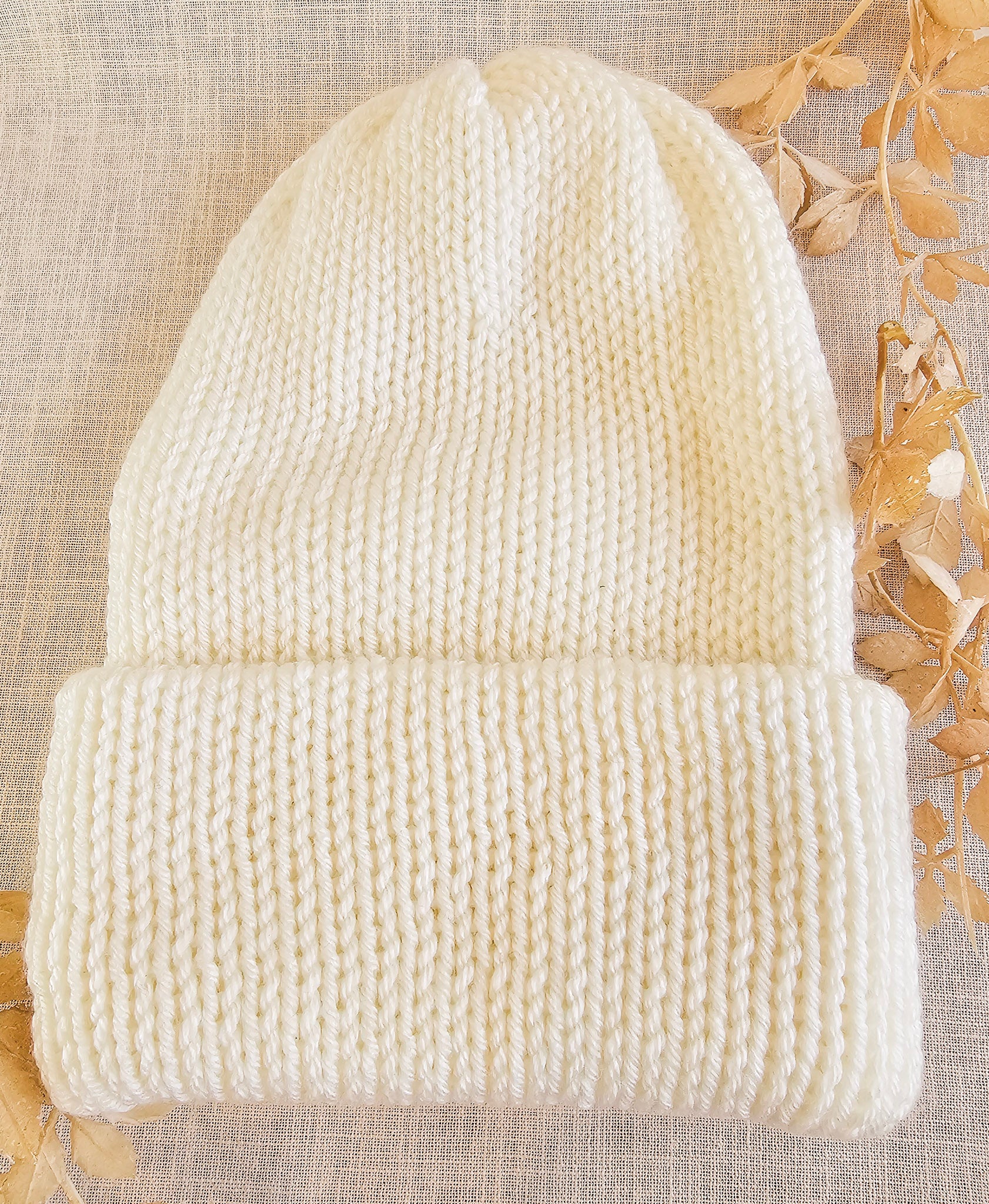 Knitted cream beanie on a textured beige background with dried leaves.