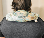 A person viewed from behind wearing a turtle-patterned hot and cold neck pack on their shoulders. The neck pack features a mosaic design in shades of teal, green, beige, and brown, with a small white fabric tag visible on the right side.
