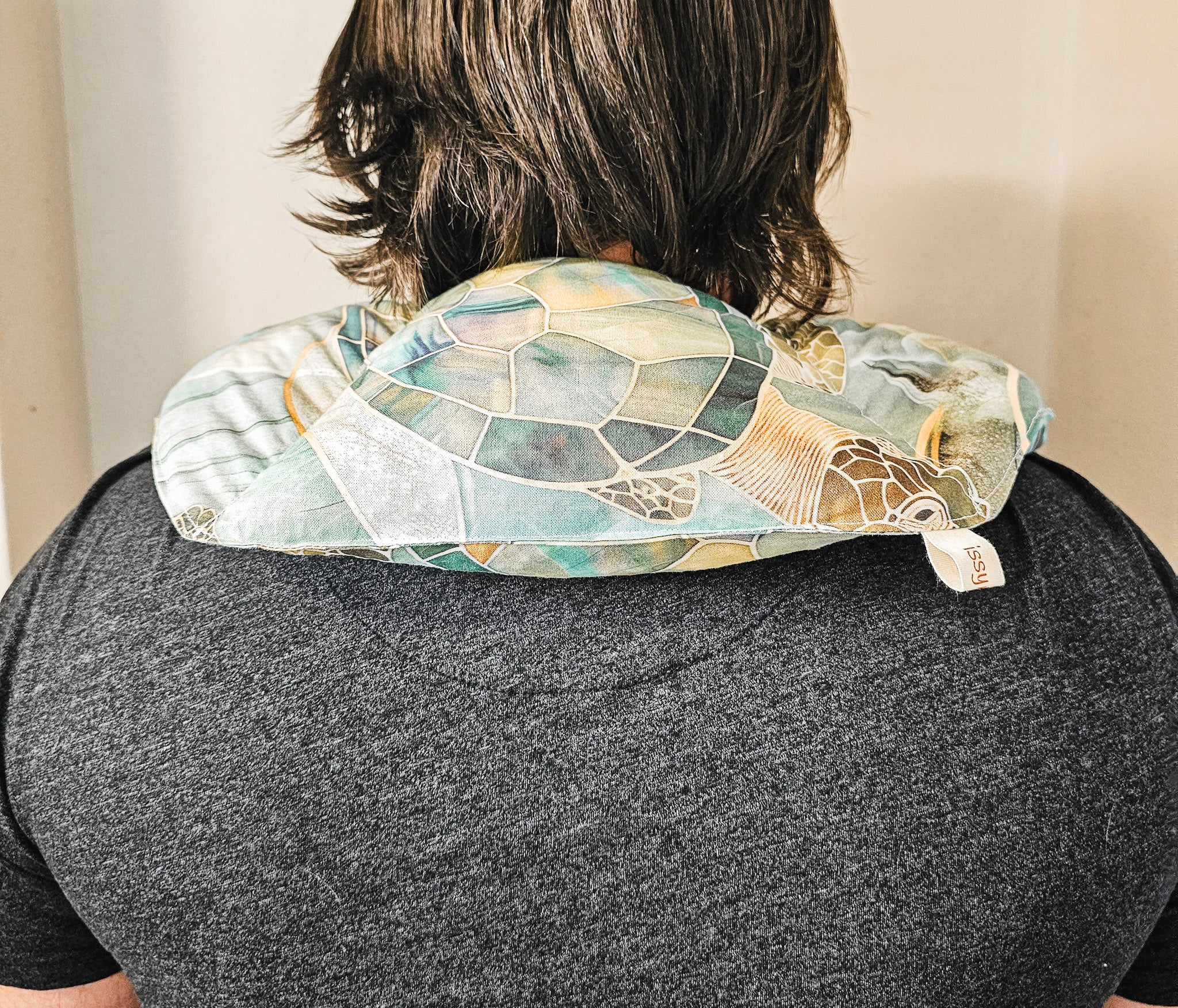 A person viewed from behind wearing a turtle-patterned hot and cold neck pack on their shoulders. The neck pack features a mosaic design in shades of teal, green, beige, and brown, with a small white fabric tag visible on the right side.