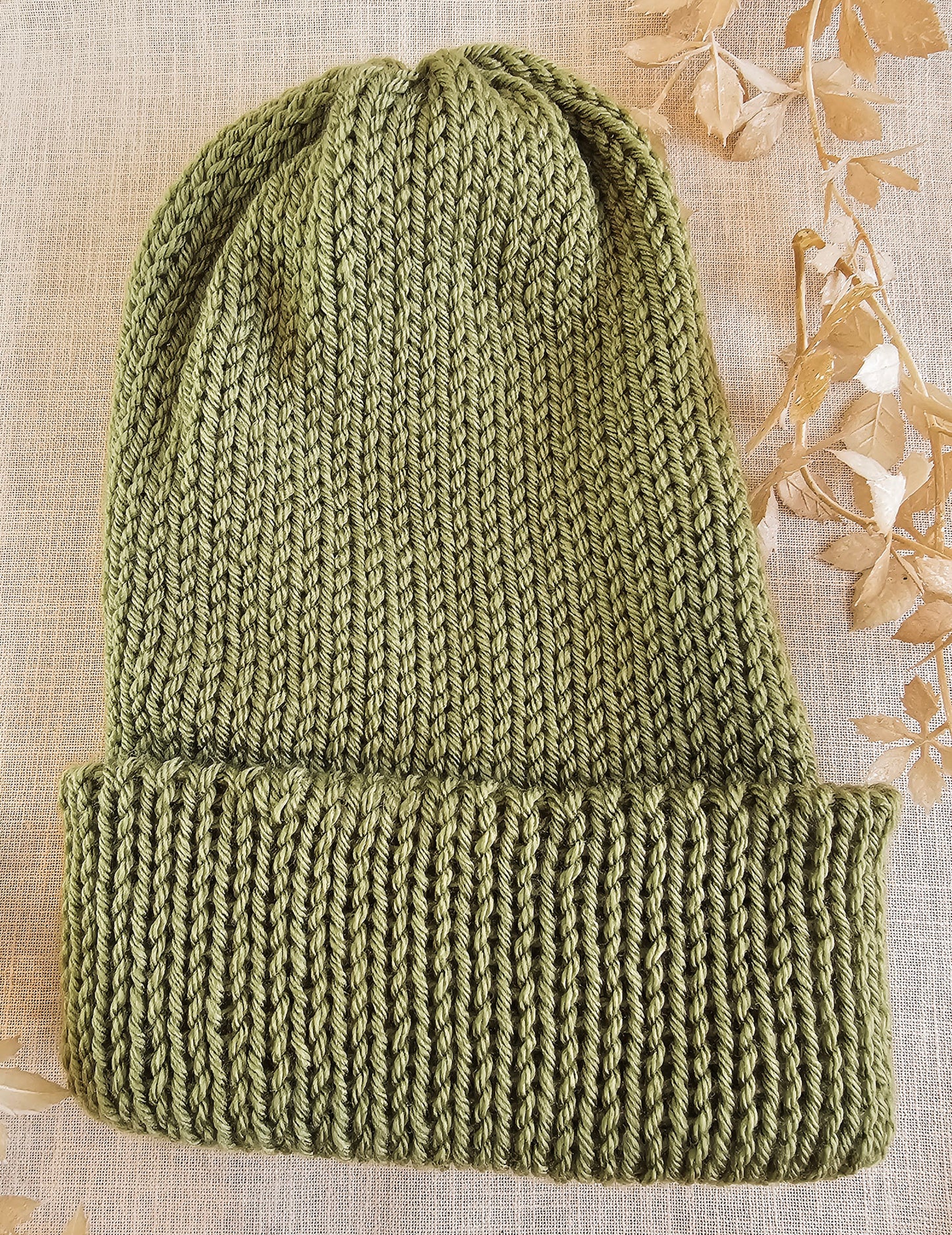 Green knitted beanie on a textured beige background with dried leaves.