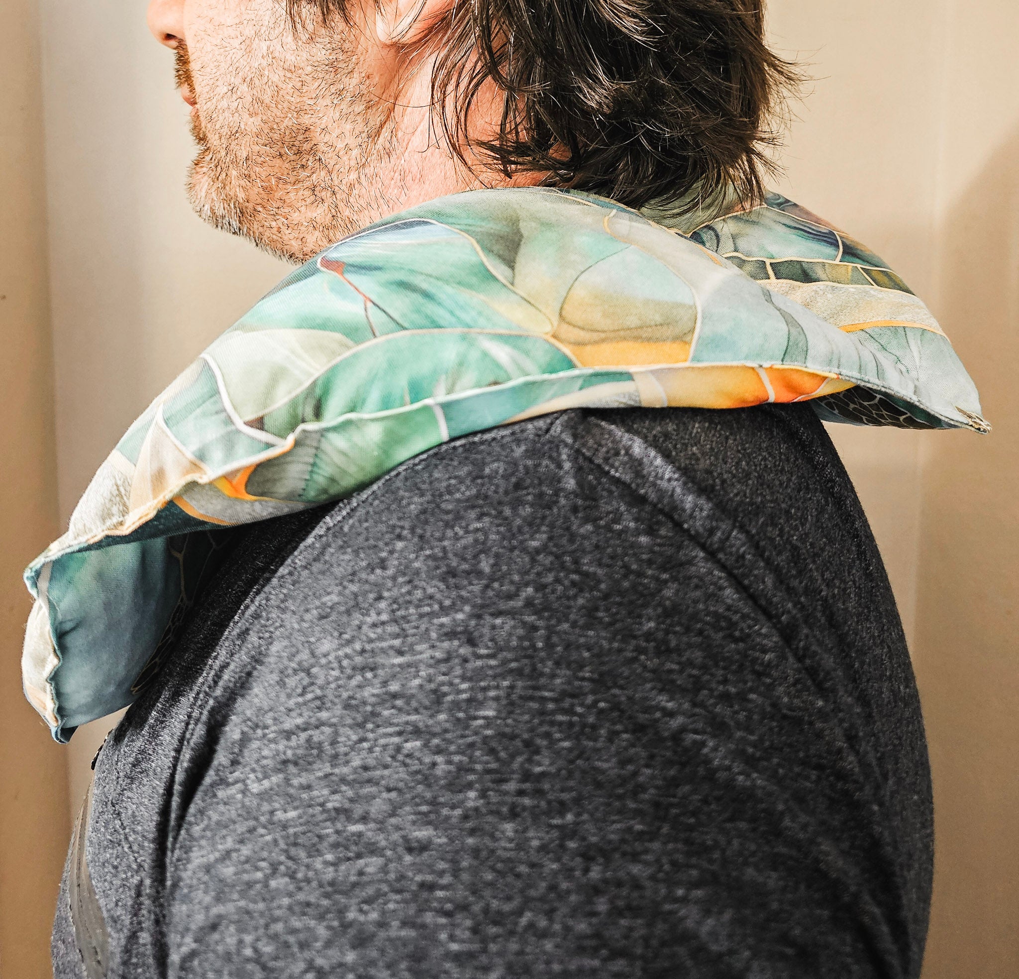 A man shown from the side wearing a patterned hot and cold neck pack with a tropical leaf design in shades of green, blue, and orange, resting on his neck over a dark gray heathered t-shirt.