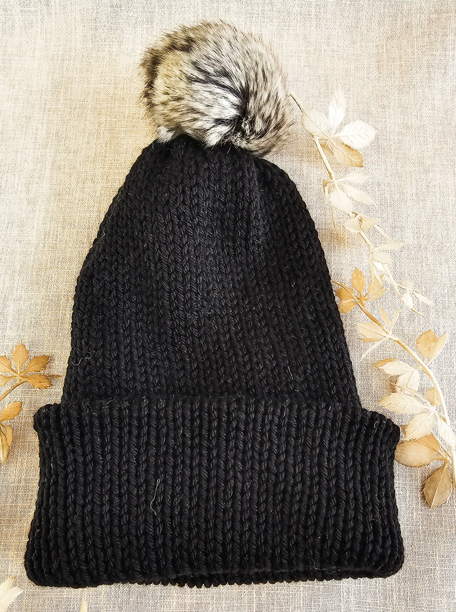 Black knit beanie with a fur pom-pom on a textured beige background