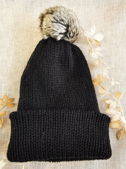 Black knit beanie with a fur pom-pom on a textured beige background