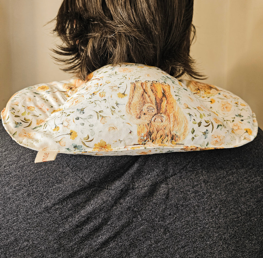 A close-up back view of a person wearing a floral-patterned neck hot and cold pack. The pillow features a cream background with yellow and green floral motifs and a central brown illustration of a tree of life.