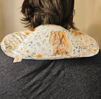 A close-up back view of a person wearing a floral-patterned neck hot and cold pack. The pillow features a cream background with yellow and green floral motifs and a central brown illustration of a tree of life.