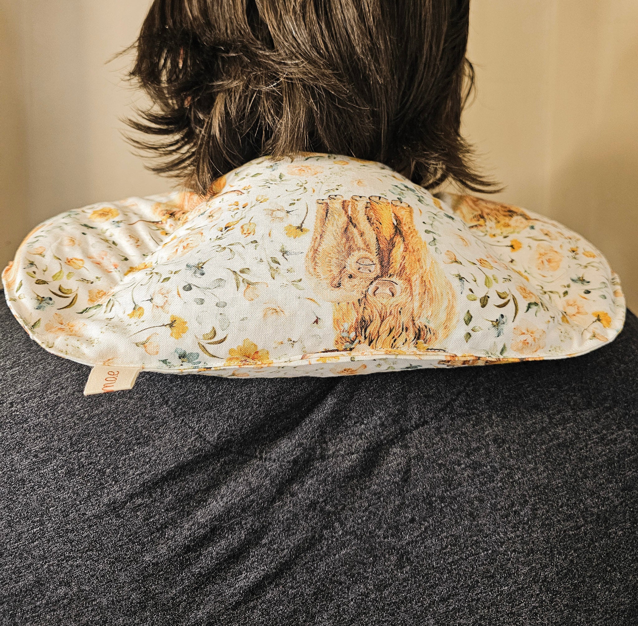 A close-up back view of a person wearing a floral-patterned neck hot and cold pack. The pillow features a cream background with yellow and green floral motifs and a central brown illustration of a tree of life.