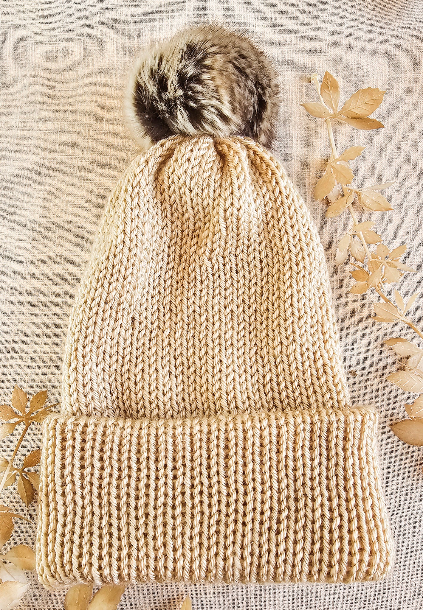 Beige knitted beanie with a fur pom-pom on a textured beige background