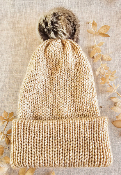 Beige knitted beanie with a fur pom-pom on a textured beige background