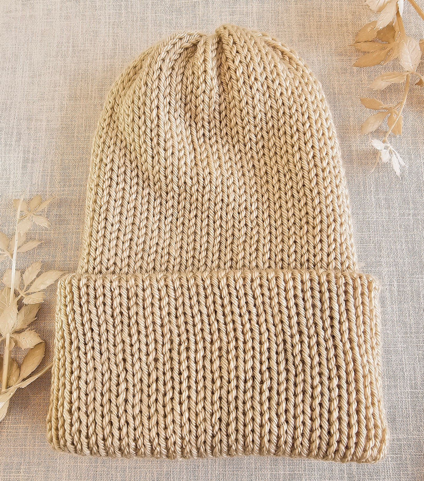 Beige knitted beanie on a textured beige background with dried leaves.