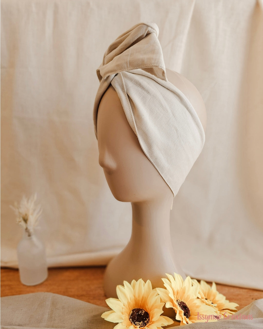 A beige cotton turban-style hair wrap displayed on a mannequin head, with dried flowers and yellow sunflowers in the foreground against a draped cream fabric backdrop.