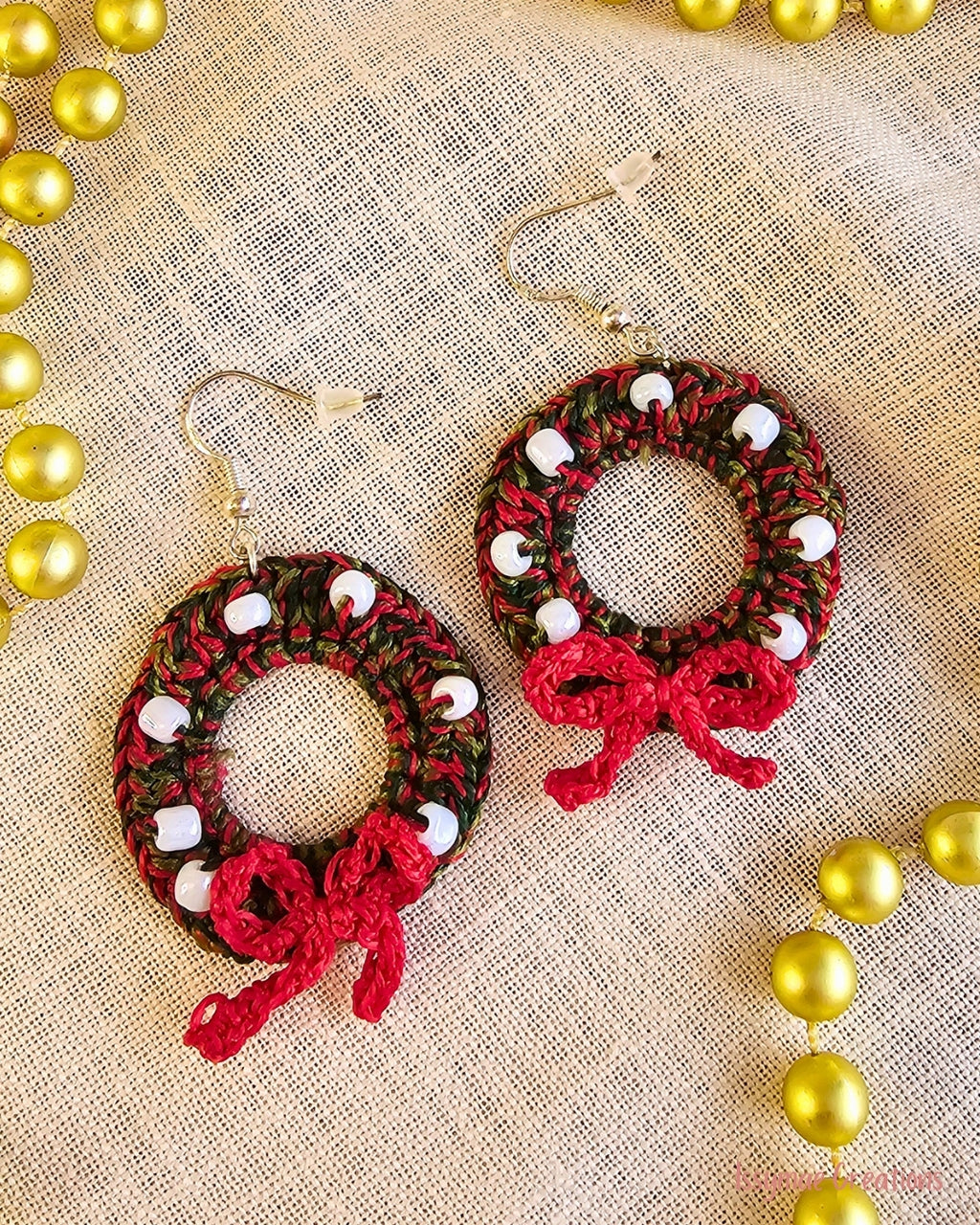 Deck the Halls crochet Christmas wreath earrings with white beads and red bows, laid flat on cream fabric.