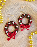 Deck the Halls crochet Christmas wreath earrings with white beads and red bows, laid flat on cream fabric.