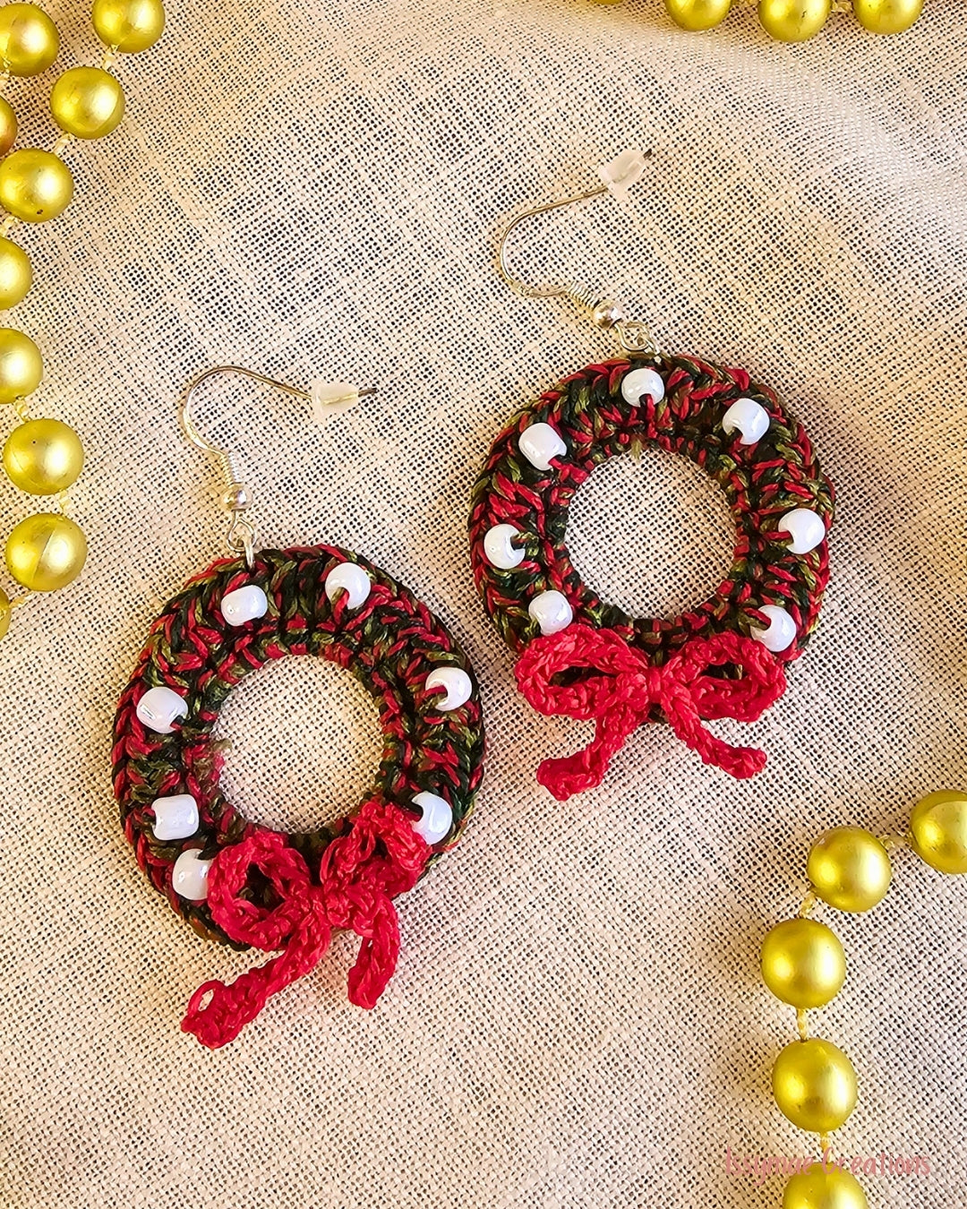 Deck the Halls crochet Christmas wreath earrings with white beads and red bows, laid flat on cream fabric.
