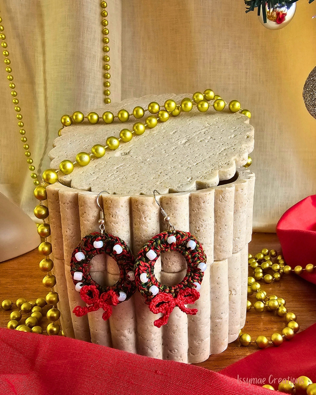Crochet Christmas wreath earrings in red and green with white beads and red bows, hanging against a cream display.