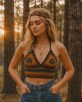 Woman wearing a colorful crochet top and denim pants in a forest setting with sunset light.