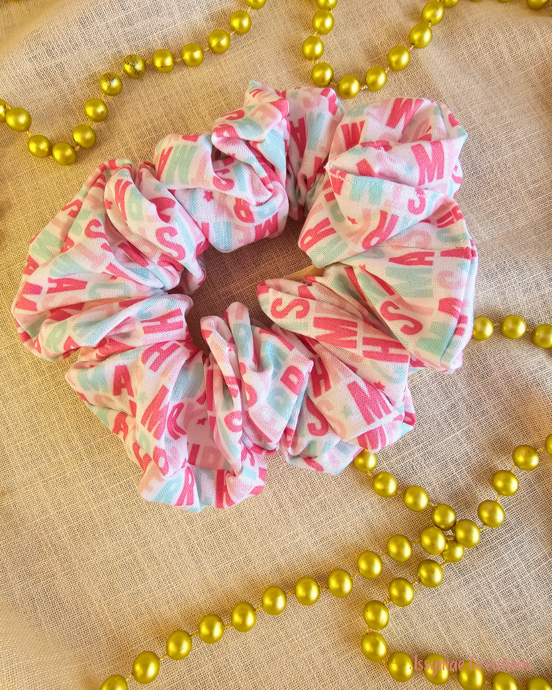 Merry & Bright Christmas scrunchie with pink lettering and pale mint checks, laid flat on cream fabric beside gold beads.