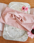 A dusty pink fabric headband or hair wrap is draped over a round marble tray, styled with pink flowers on a wooden surface.