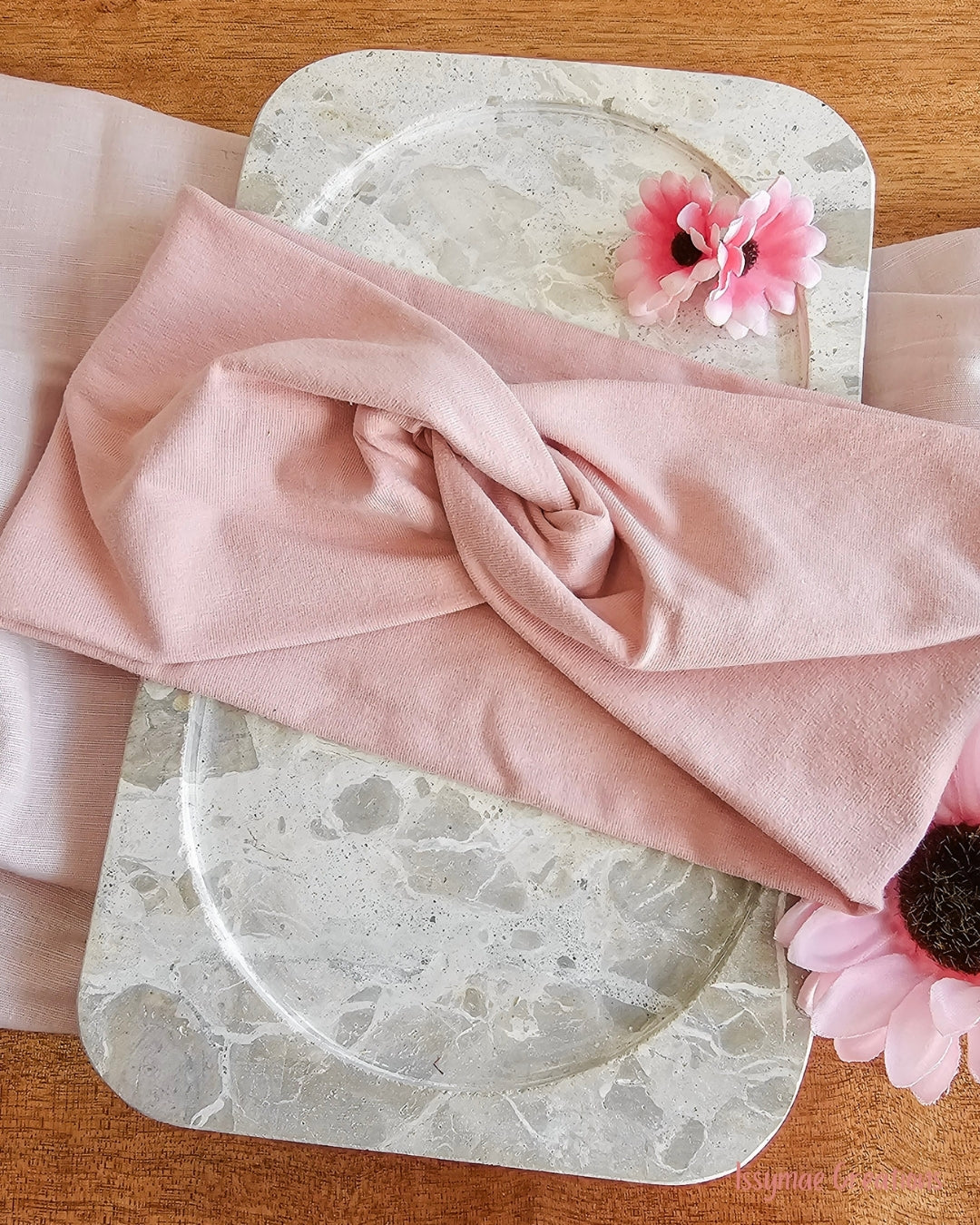 A dusty pink fabric headband or hair wrap is draped over a round marble tray, styled with pink flowers on a wooden surface.
