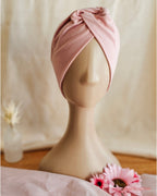 A dusty pink fabric headwrap styled on a beige mannequin head, set against a soft beige fabric backdrop with decorative pink flowers in the foreground.