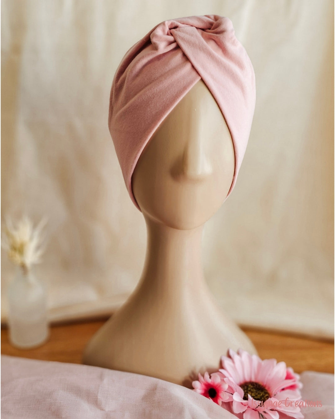 A dusty pink fabric headwrap styled on a beige mannequin head, set against a soft beige fabric backdrop with decorative pink flowers in the foreground.