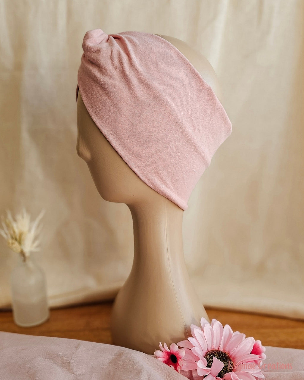 A light pink fabric head wrap or turban-style hair tie displayed on a beige mannequin head, with soft neutral drapery in the background and pink flowers at the base.