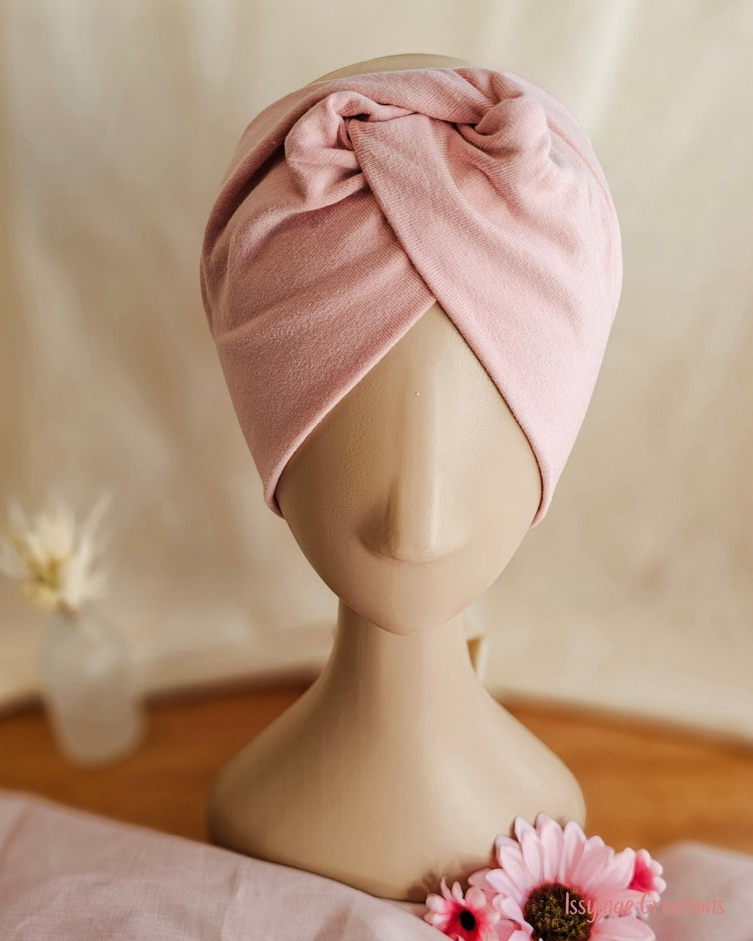 A light pink cotton turban wrap headband displayed on a beige mannequin head, set against a soft beige fabric backdrop with decorative flowers.