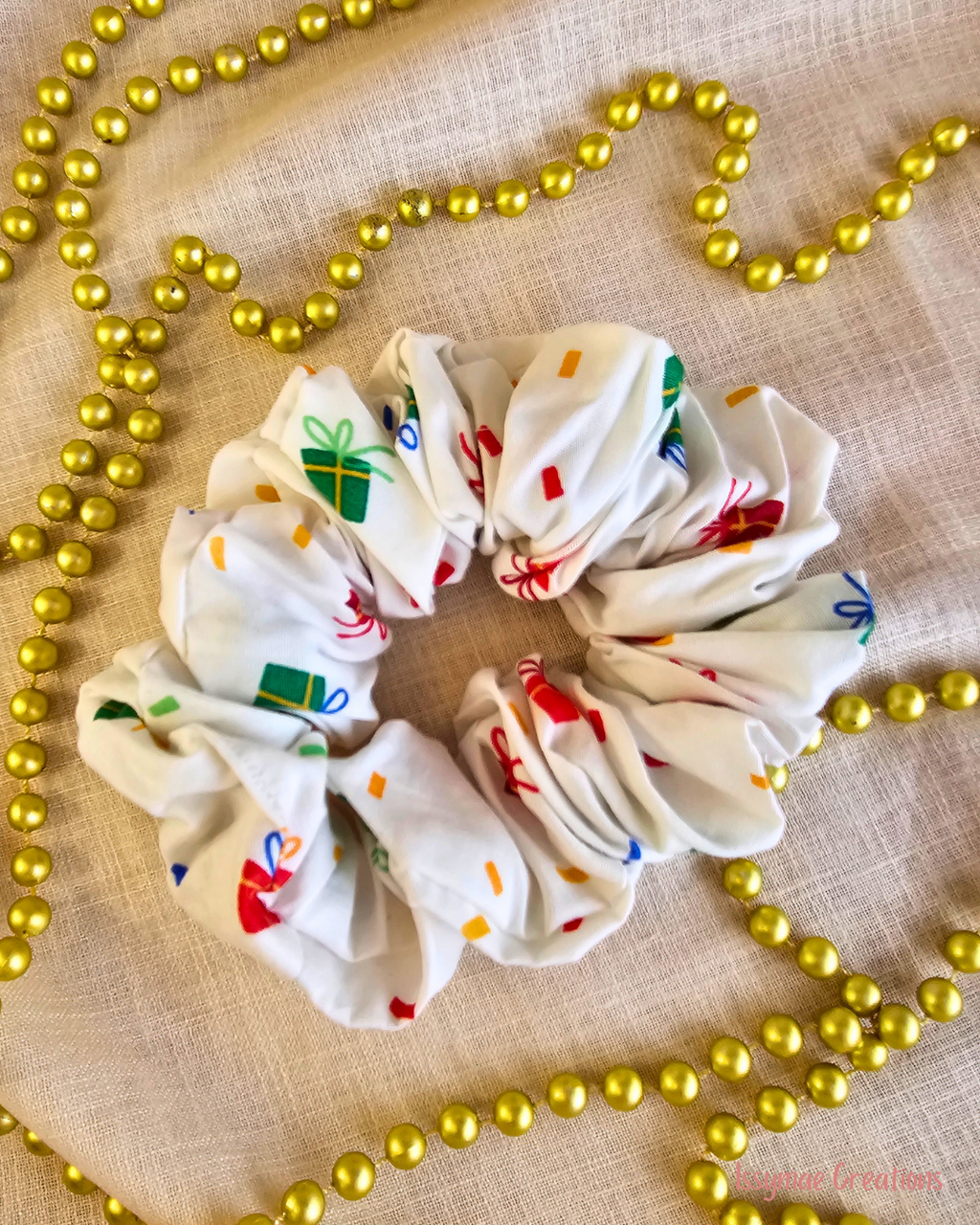 Present Time white Christmas scrunchie with colourful wrapped gift prints, laid flat on cream fabric beside gold beads.