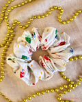 Present Time white Christmas scrunchie with colourful wrapped gift prints, laid flat on cream fabric beside gold beads.