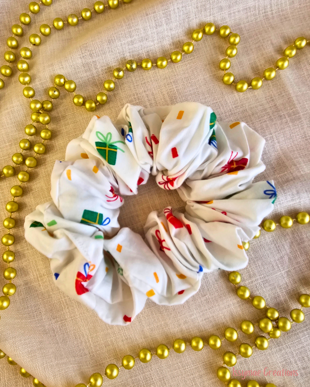 Present Time white Christmas scrunchie with colourful wrapped gift prints, laid flat on cream fabric beside gold beads.