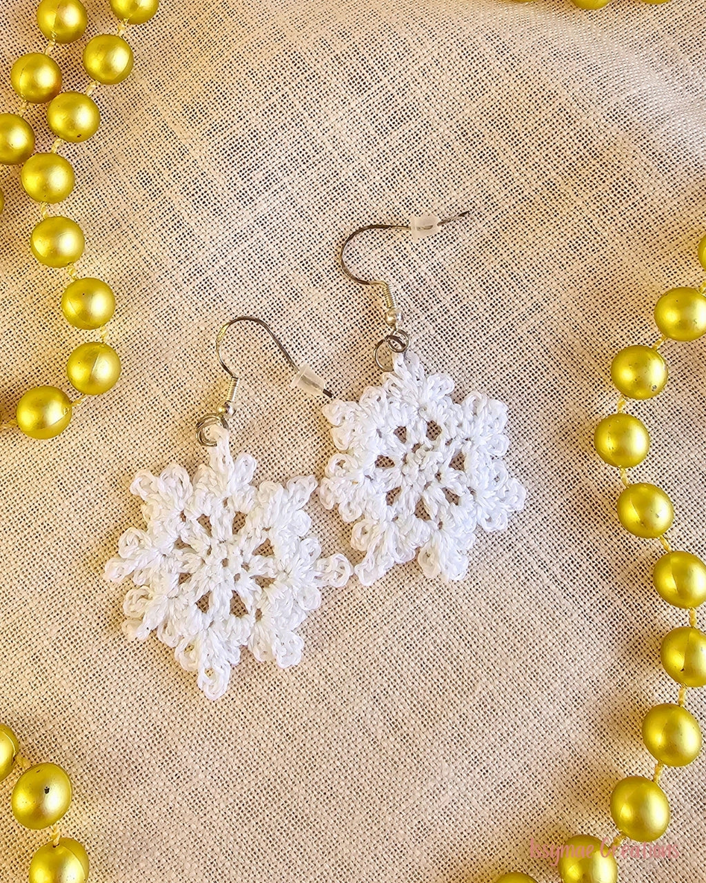 Snow Much Fun white crochet snowflake earrings laid flat on cream fabric beside gold Christmas garland.