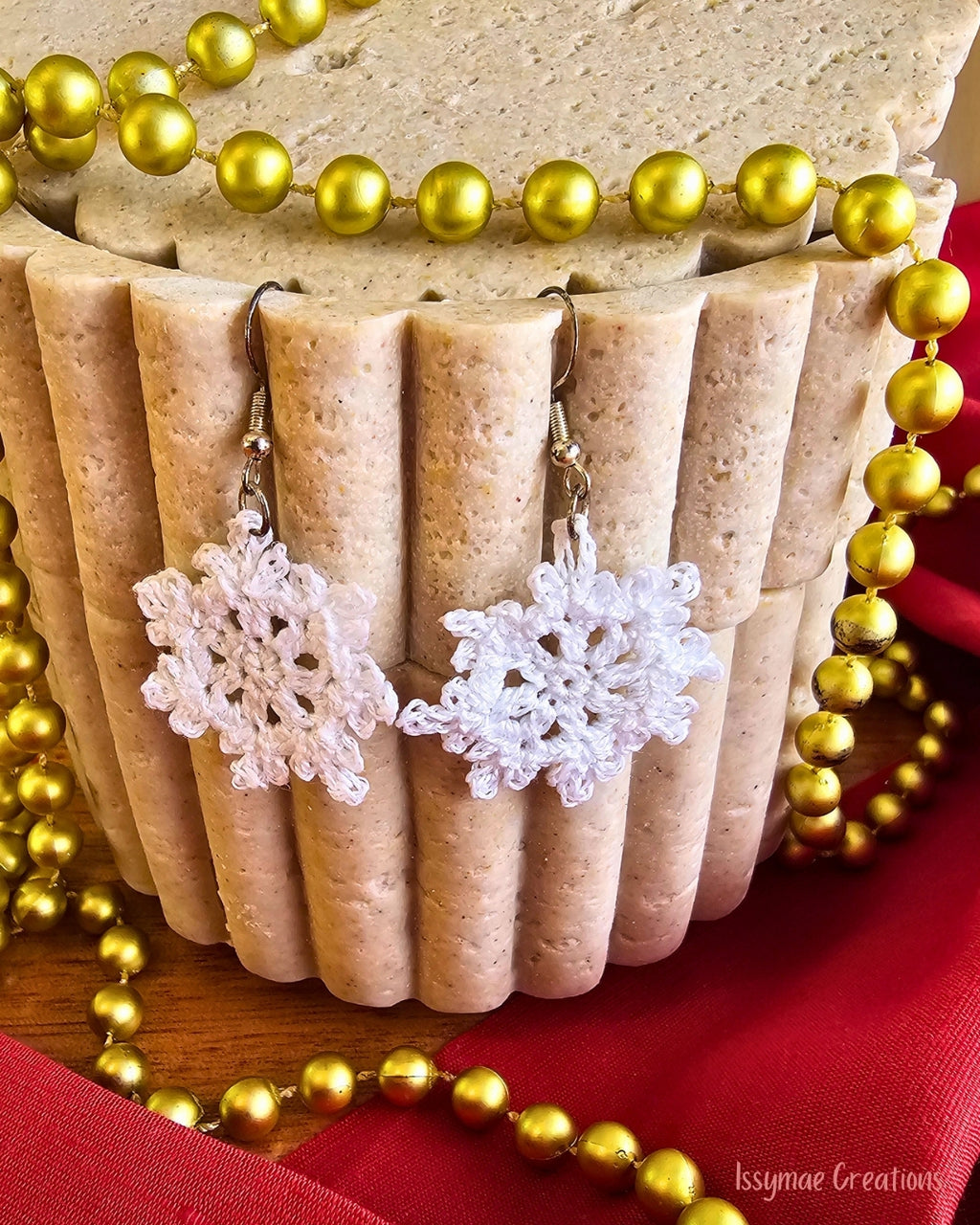 White crochet snowflake earrings hanging against a cream fluted display, surrounded by gold Christmas beads.