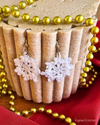 White crochet snowflake earrings hanging against a cream fluted display, surrounded by gold Christmas beads.
