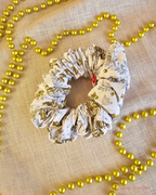 Frosted Holly white Christmas scrunchie with gold and silver holly print, laid flat on cream fabric beside gold beads.