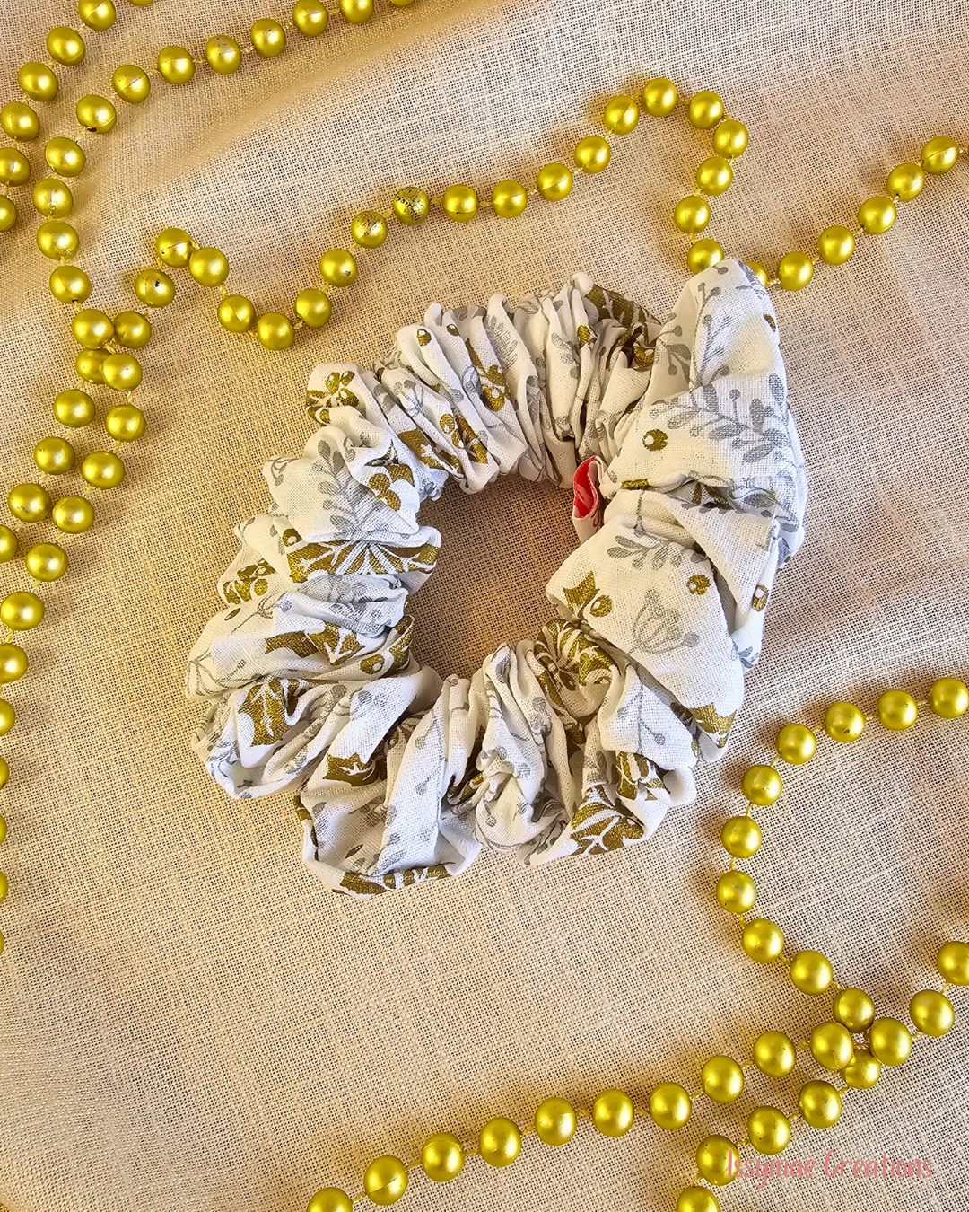 Frosted Holly white Christmas scrunchie with gold and silver holly print, laid flat on cream fabric beside gold beads.