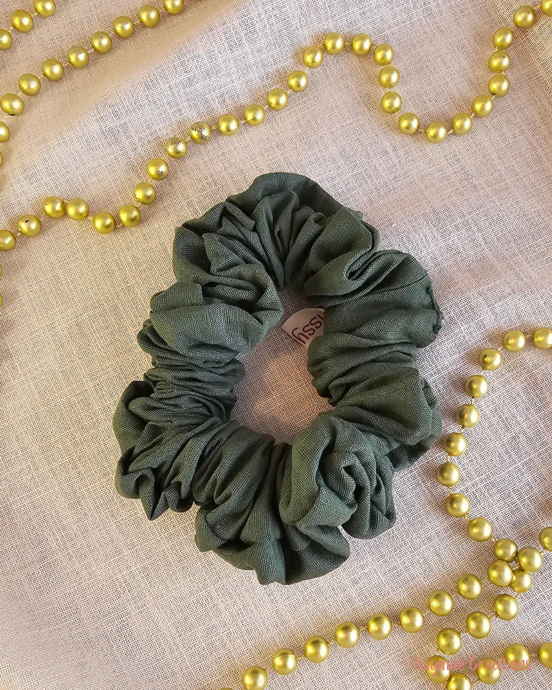 An olive green fabric scrunchie with a gathered, ruffled texture, displayed on a beige linen fabric background with decorative gold pearl beads.