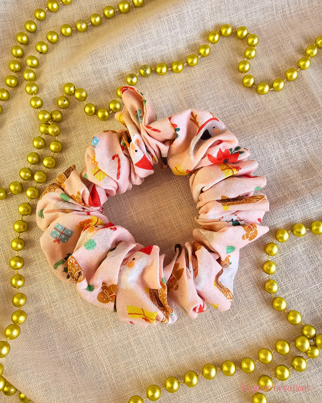 Gingerbread Gang pink Christmas scrunchie with gingerbread figures, Santa faces and gifts, laid flat beside gold beads.