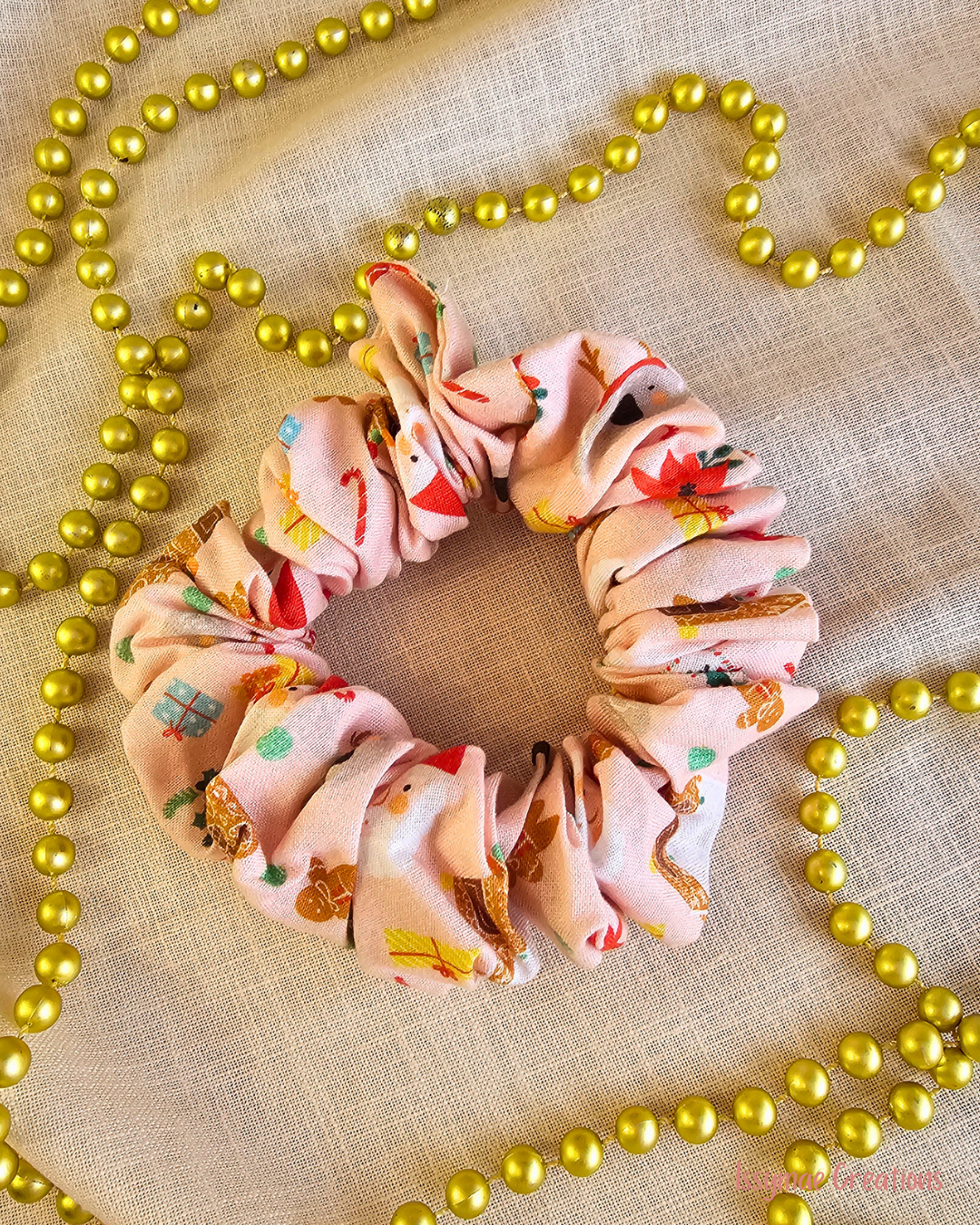 Gingerbread Gang pink Christmas scrunchie with gingerbread figures, Santa faces and gifts, laid flat beside gold beads.