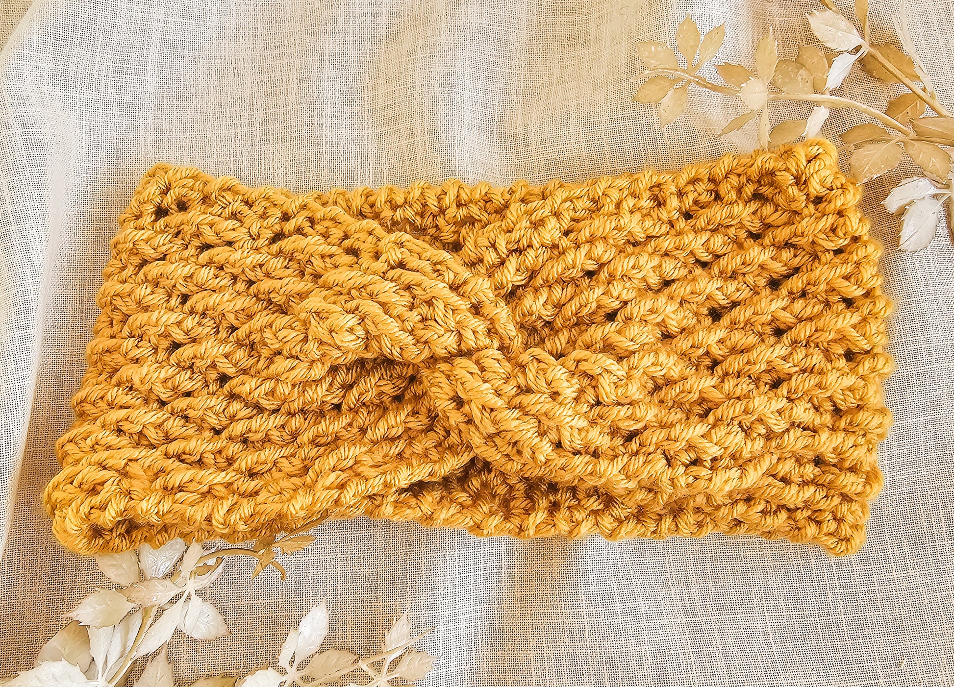 Mustard yellow knitted headband on a textured beige background with floral elements.